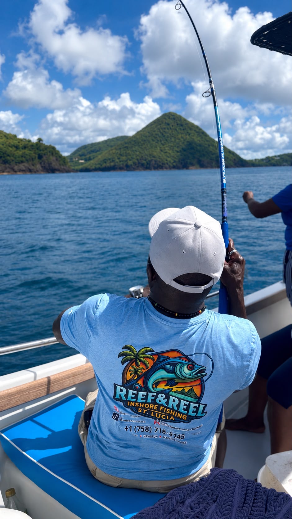 A Reef & Reel guide in a branded T-shirt reeling in a fish off the Saint Lucia coast, with a green hill and blue sea ahead